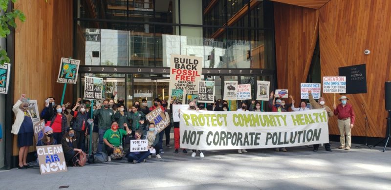 Community members in favor of Rule 6-5 gather for a group picture in front of BAAQMD's headquarters in San Francisco with several signs. One major banner reads, " protect community health, not corporate wealth"